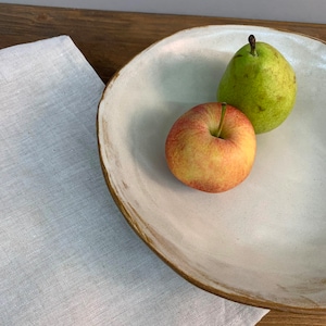 May include: A white ceramic serving bowl with a brown rim, featuring a green pear and a red apple on a white linen tablecloth.