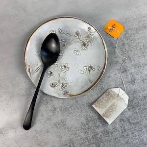 May include: A black spoon rests on a white ceramic spoon rest with a floral design and a gold rim. A tea bag with an orange tag is next to the spoon rest. The items are on a gray surface.