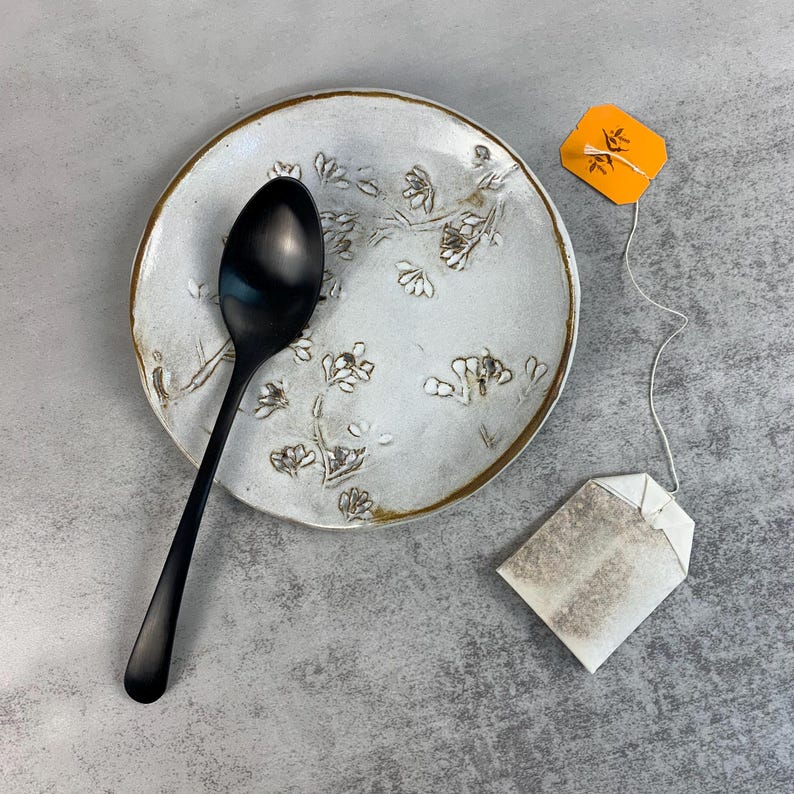 May include: A black spoon rests on a white ceramic spoon rest with a floral design and a gold rim. A tea bag with an orange tag is next to the spoon rest. The items are on a gray surface.