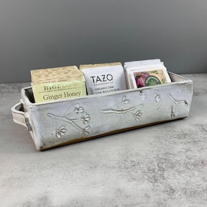 May include: A rectangular, off-white ceramic tea bag holder with embossed floral designs. The holder contains tea bags from Bigelow Ginger Honey, Tazo Organic Chai, and Traditional Medicinals. The holder has two small handles on the sides.