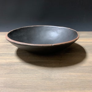 May include: A black ceramic bowl with a slightly raised rim. The bowl is sitting on a wooden surface.