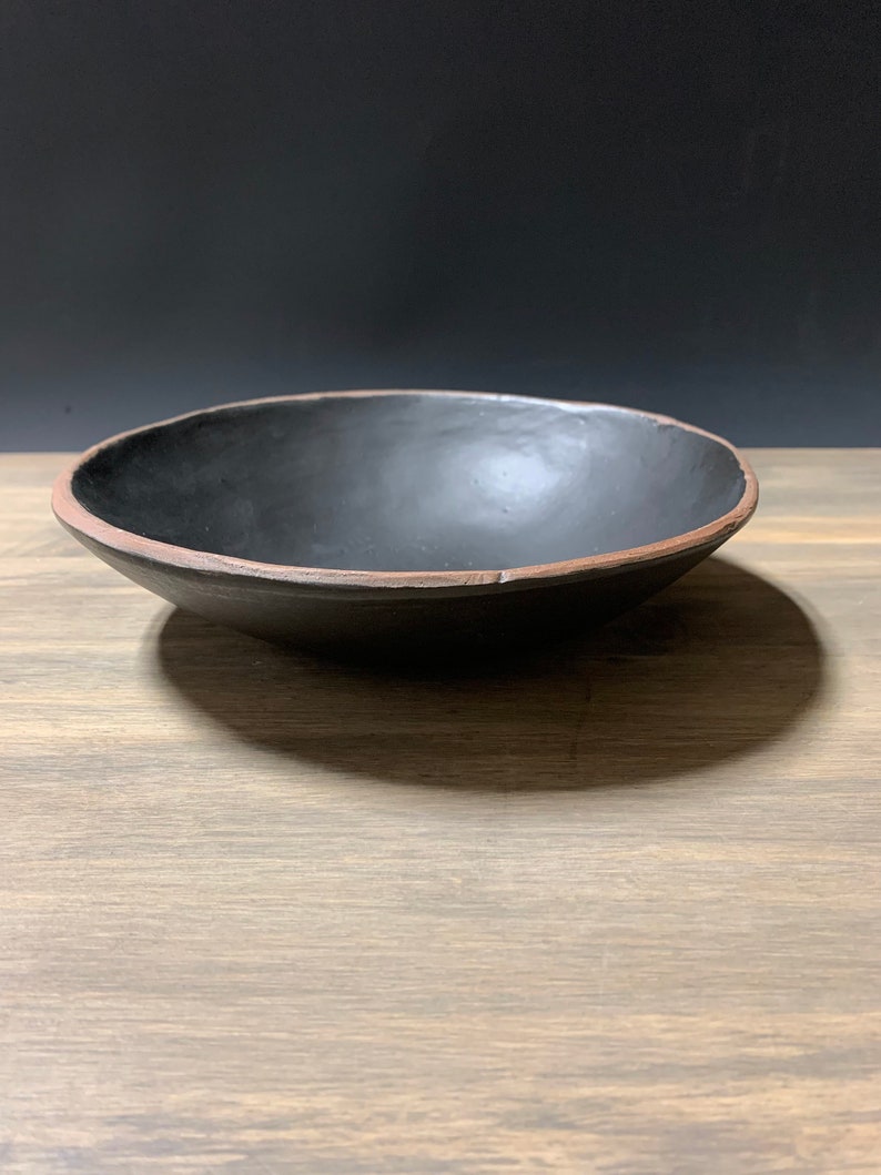 May include: A black ceramic bowl with a slightly raised rim. The bowl is sitting on a wooden surface.