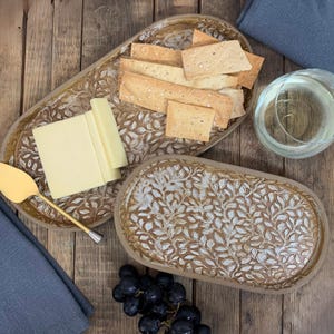 May include: Two oval serving platters with a leaf design, one holding crackers and the other cheese. A gold cheese knife, a glass of water, and a bunch of grapes are also present. The platters are on a wooden surface.