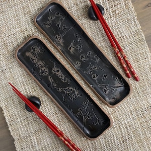 May include: Two rectangular black ceramic chopstick rests with floral designs and a brown rim. Two pairs of red chopsticks with gold floral accents rest on the chopstick rests and black chopstick holders. The items are on a beige woven placemat.