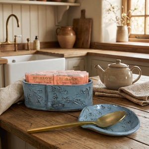 May include: A blue ceramic tea bag holder with floral relief, filled with Bigelow Ginger Peach tea bags. A matching spoon rest holds a gold spoon. A beige teapot sits nearby on a wooden table, creating a cozy kitchen scene.