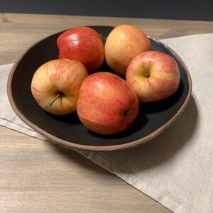 May include: Five red apples in a black bowl with a brown rim. The bowl is sitting on a light brown wooden surface with a beige linen napkin underneath.
