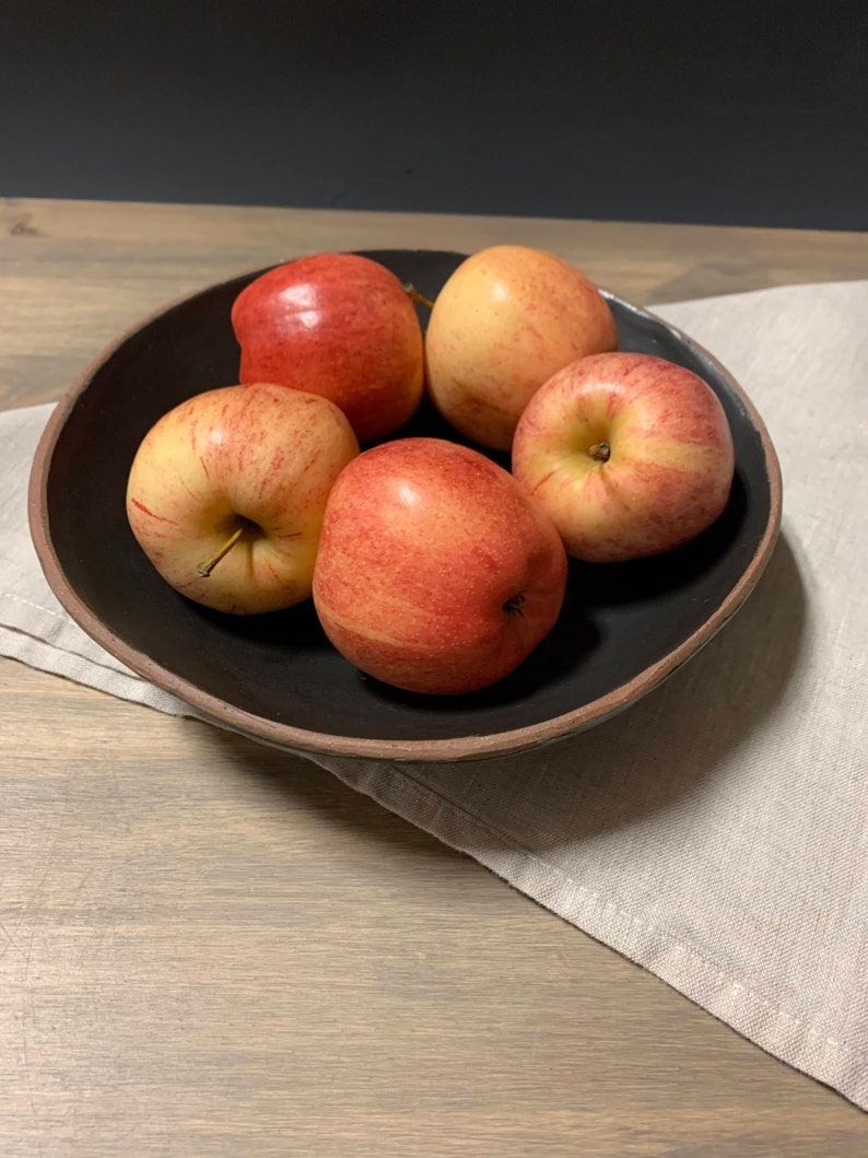 May include: Five red apples in a black bowl with a brown rim. The bowl is sitting on a light brown wooden surface with a beige linen napkin underneath.