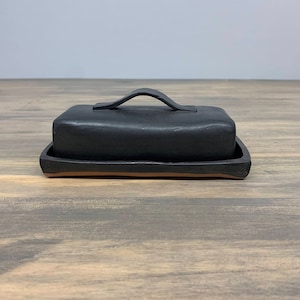 May include: Black ceramic butter dish with a handle. The dish has a rectangular shape and a smooth, matte finish.