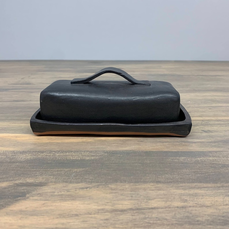 May include: Black ceramic butter dish with a handle. The dish has a rectangular shape and a smooth, matte finish.