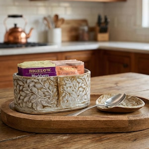 May include: A wooden serving tray holds a decorative ceramic tea caddy with leaf patterns, tea packets, a small dish, and a spoon. The tea caddy contains Bigelow Blackberry and Ginger Peach tea. The tray sits on a wooden table.