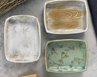 May include: Three ceramic soap dishes with different designs. The first dish is white with a raised leaf pattern. The second dish is white with a wood grain pattern. The third dish is green with a floral pattern.