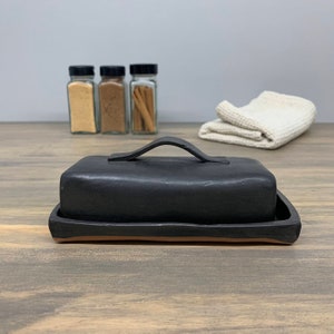 May include: Black ceramic butter dish with a handle and a terracotta rim. The dish is sitting on a wooden surface.