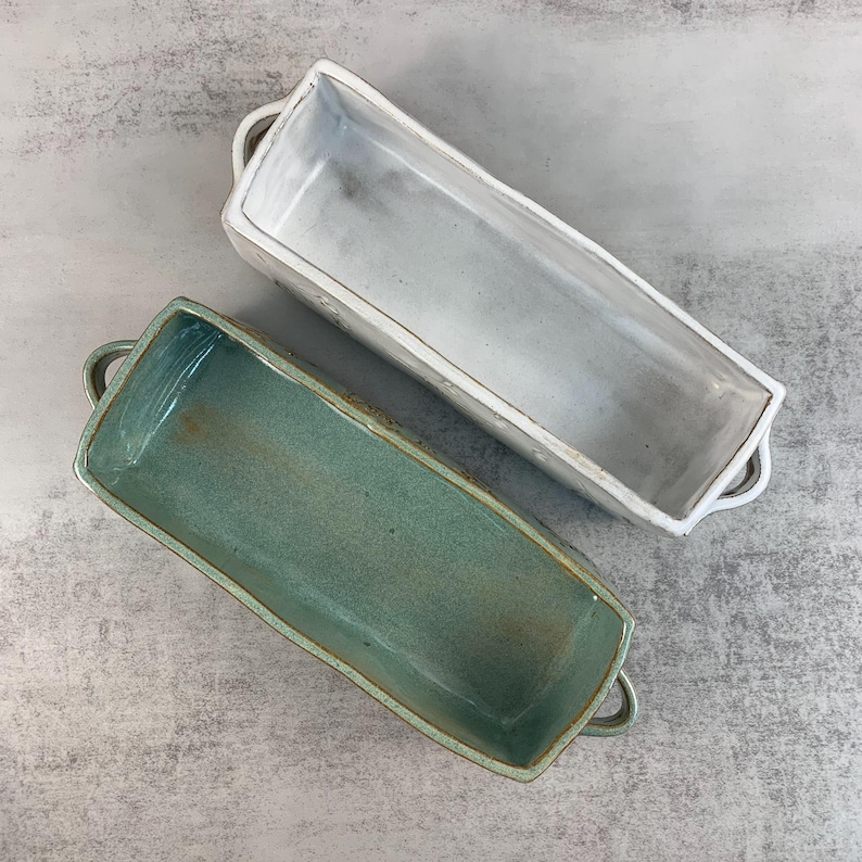 May include: Two rectangular ceramic baking dishes. One is a light gray color, and the other is a teal color. Both have small handles on each end. The dishes are empty and sit on a gray surface.
