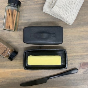 May include: A black ceramic butter dish with a lid and a stick of butter inside. The dish is on a wooden surface.