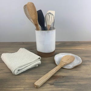 May include: White ceramic utensil holder with a brown rim, filled with wooden spoons and a whisk. A matching white ceramic spoon rest with a wooden spoon on top sits on a wooden surface.