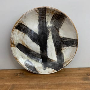 May include: A handmade ceramic bowl with a white interior and a brown rim. Bold black brushstrokes cross the white surface, creating an abstract design. The bowl sits on a wooden surface.