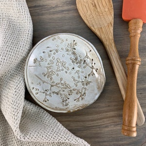 May include: A white ceramic spoon rest with a floral design. The rest is handmade and has a rustic, farmhouse style.