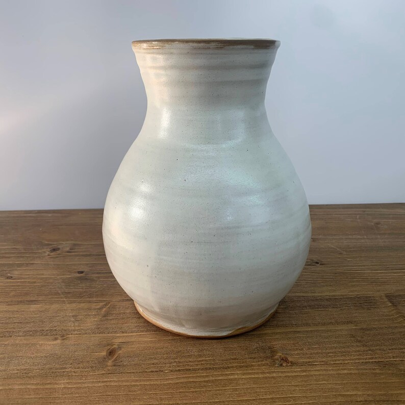 May include: A white ceramic vase with a rounded body and a slightly flared opening. The vase has a light brown rim and a subtle glaze.
