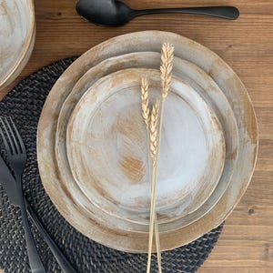 May include: A rustic table setting featuring a stack of three off-white plates with a textured, brushed design. A black spoon, fork, and knife are arranged on a dark gray woven placemat. Wheat stalks add a natural touch to the arrangement.