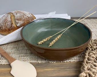 Puede incluir: Un cuenco de cerámica hecho a mano con interior verde y exterior marrón. Dos espigas de trigo descansan dentro del cuenco. Un pan y una espátula también están en la imagen, lo que sugiere un tema de cocina o comedor.