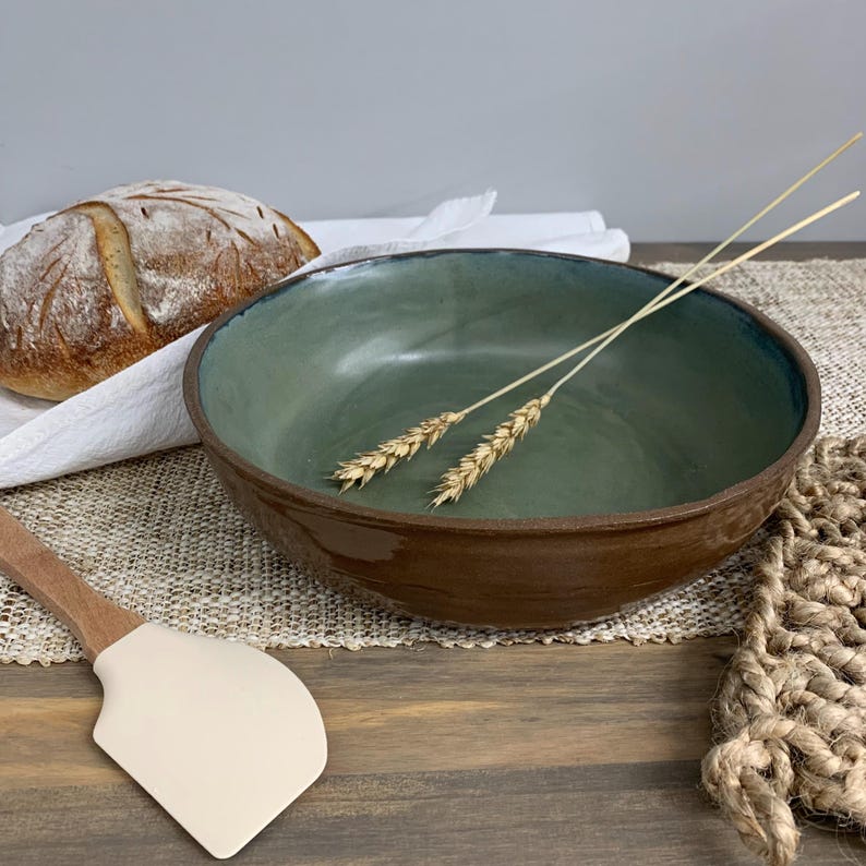 May include: A handmade ceramic bowl with a green interior and brown exterior. Two stalks of wheat rest inside the bowl. A loaf of bread and a spatula are also in the image, suggesting a kitchen or dining theme.
