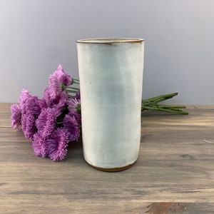 May include: A white ceramic vase with a brown rim. The vase is cylindrical and has a smooth, matte finish. It is sitting on a wooden surface with a bouquet of purple flowers.