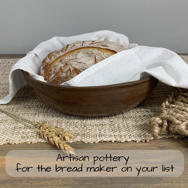 May include: A brown ceramic bread bowl holding a loaf of bread covered with a white cloth. The bowl sits on a woven mat with a sprig of wheat. Text at the bottom reads: Artisan pottery for the bread maker on your list.