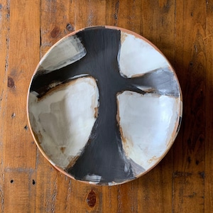 May include: A round ceramic serving bowl with a hand-painted abstract design. The bowl features a black, white, and tan color palette. The interior has a black central shape with white and tan accents. The rim is a light tan color.