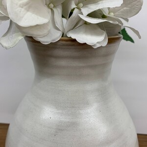 May include: White ceramic vase with a wide mouth and a rounded body. The vase has a subtle speckled texture and a light brown rim.