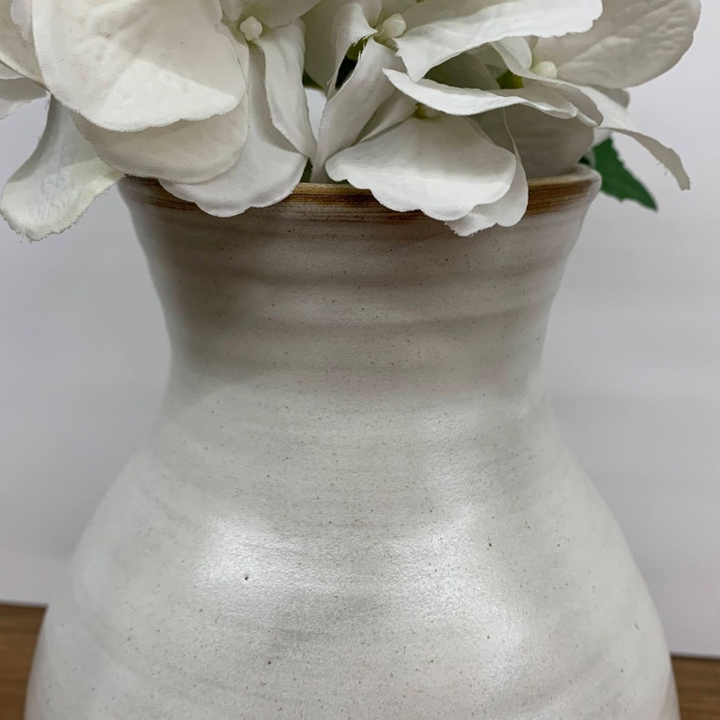 May include: White ceramic vase with a wide mouth and a rounded body. The vase has a subtle speckled texture and a light brown rim.