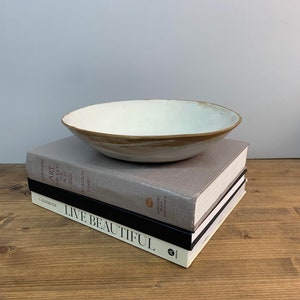 Puede incluir: Un cuenco de cer&aacute;mica blanco con un borde marr&oacute;n se encuentra sobre una pila de libros. Los libros tienen t&iacute;tulos como "Art Through the Ages", "De La Croix Tansey", "Live Beautiful" y "Harcourt Brace Jovanovich".
