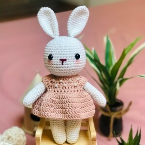 May include: A white crocheted bunny wearing a pink dress with pearl accents. The bunny is sitting on a wooden stool.