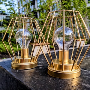 May include: Two gold metal cage lanterns with light bulbs inside. The lanterns are sitting on a black surface.