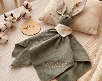 Peut inclure: Une couverture de sécurité en mousseline vert sauge avec une tête de lapin en peluche aux longues oreilles, brodée du nom Nelio. Les oreilles du lapin ont une doublure en tissu blanc avec un petit motif animalier marron. Un disque en bois affiche Tani.