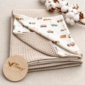 Könnte beinhalten: Eine gefaltete Waffelpiqué-Decke in hellem Beige. Das Innenfutter besteht aus weißem Stoff mit einem wiederkehrenden Muster aus braunen und grünen Baufahrzeugen, darunter Muldenkipper, Traktoren und Kräne. Ein rundes Holzschild trägt die Aufschrift Tavi.