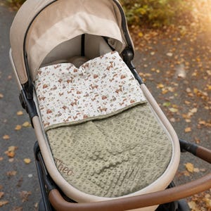 Op de afbeelding: Een kinderwagen met een beige kap en een tweekleurige deken. De deken heeft een wit bovendeel met een bruin en groen bosdierenpatroon en een saliegroen, genopt minky-onderdeel dat is geborduurd met de naam Theo.