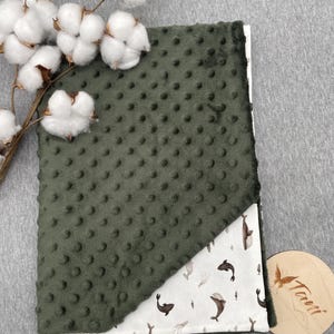 May include: A green, textured baby blanket with a white corner featuring a whale pattern. The blanket is made of soft material and has a dotted texture. A sprig of cotton adds a decorative touch.