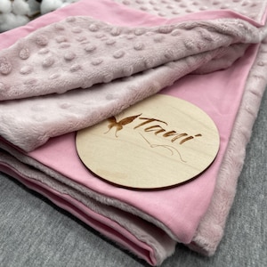 Może przedstawiać: Różowo-biały kocyk dla niemowląt o miękkiej, pluszowej fakturze. Kocyk jest złożony na pół i ma drewniane kółko z wygrawerowanym napisem "Tavi".