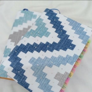 May include: Two notebooks with a white background. The covers have a blue, grey, and white zigzag pattern with small, scattered, pearl-like embellishments. The notebooks have a blue ribbon and colourful page dividers.