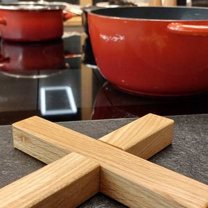 Può includere: Un sottopentola in legno a forma di X, realizzato in legno marrone chiaro. Il sottopentola è appoggiato su un piano di lavoro grigio scuro.