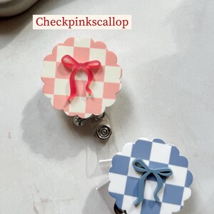 Pu&ograve; includere: Due porta badge decorativi. Uno &egrave; a scacchi rosa e bianchi con un fiocco rosa, l'altro &egrave; a scacchi blu e bianchi con un fiocco blu. Il testo "Checkpinkscallop" e "Dustycheckscallop" &egrave; stampato sull'immagine.