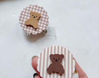 May include: Two circular badge reels with scalloped edges. One features a light brown and white gingham pattern with a tan bear figure. The other features a beige and white vertical striped pattern with a dark brown bear figure attached to the centre.