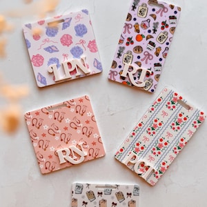 May include: Five rectangular badge holders with various patterns and initials. The patterns include seashells, food items, ballet shoes, floral designs, and books with bows. The initials are IPN, RN, PCT, and CMA. The badges are white with a hole at the top.