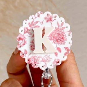 May include: A floral-themed badge reel with a white background and pink flower pattern. The letter "K" is prominently displayed in the center. The reel has a scalloped edge and a clear plastic clip.
