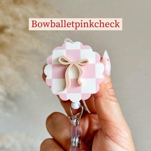Pu&ograve; includere: Un portabadge a scacchi rosa e bianchi con un design a fiocco. Il portabadge ha un bordo smerlato e una clip di plastica trasparente. Il testo "Bowballetpinkcheck" &egrave; visualizzato sopra il portabadge.