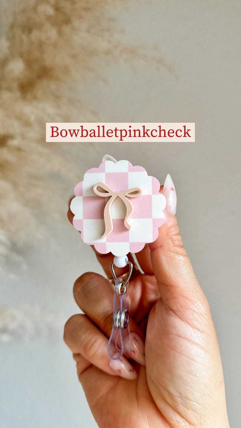 Pu&ograve; includere: Un portabadge a scacchi rosa e bianchi con un design a fiocco. Il portabadge ha un bordo smerlato e una clip di plastica trasparente. Il testo "Bowballetpinkcheck" &egrave; visualizzato sopra il portabadge.