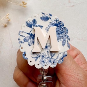 May include: White badge reel with a blue and white floral pattern and a white letter "M" in the center.