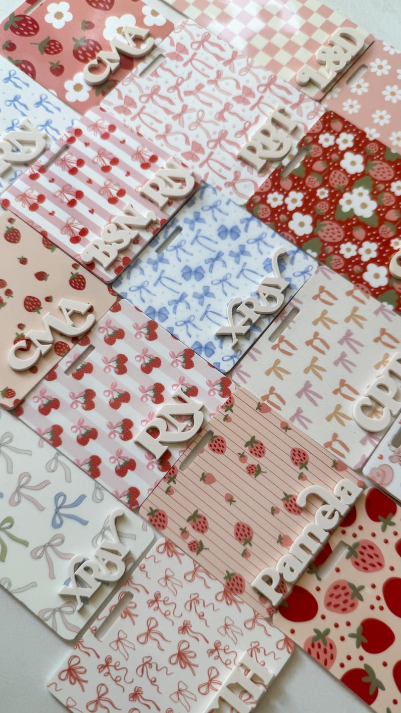 May include: A collection of decorative name tags with various patterns, including strawberries, cherries, bows, and gingham. Each tag has a white, raised name. The color palette includes pink, red, and white.