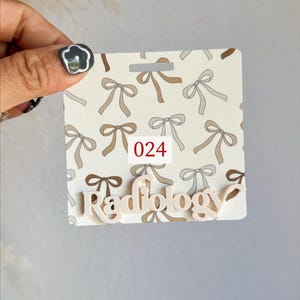 May include: A badge with a bow pattern in shades of brown and gray. The badge has the word "Radiology" in a light color and the number "024" in a red box.
