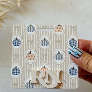 May include: A square badge reel cover featuring a beige and white vertical striped background with a repeating pattern of pumpkins in scalloped oval frames and small bows. The white letters RN are attached to the front of the badge reel cover.
